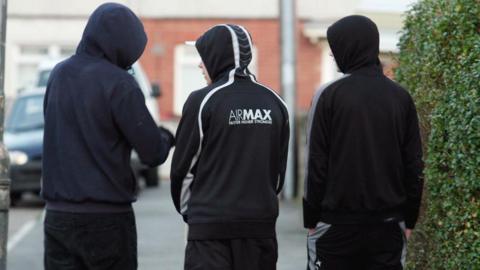 Three men, dressed in black hoodies and trousers, have their backs turned. They are walking in a neighbourhood. 
