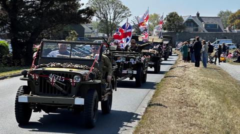 The image shows a procession of military vehicles decorated with flags, driving along a road by a grass bank with people watching.