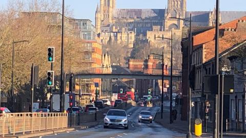 A city centre scene, which shows a busy road, with traffic heading towards the centre of the picture. Above is a pedestrian bridge, which crosses the road, with Lincoln Cathedral perched high in the background. 