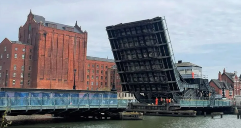 The bridge in its lifted position with men in orange suits underneath surveying the steel work.  Old red brick buildings of the original dock stand proud behind it.