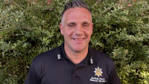 Mark Cowling is smiling and looking at the camera. He is wearing a shirt with the Suffolk Fire and Rescue Service badge and logo. 