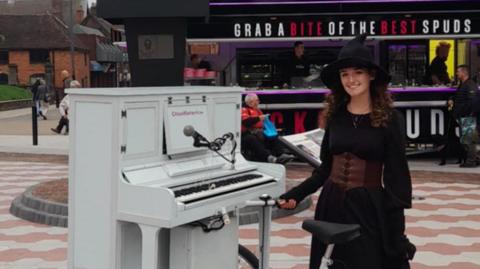 Ms Aston is wearing a witch outfit and is standing next to a white piano on a bike. A microphone is attached to the instrument. She is standing outside Spudman in Tamworth.