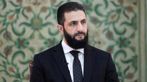 Ahmed al-Sharaa sits in a suit and tie in front of patterned wallpaper