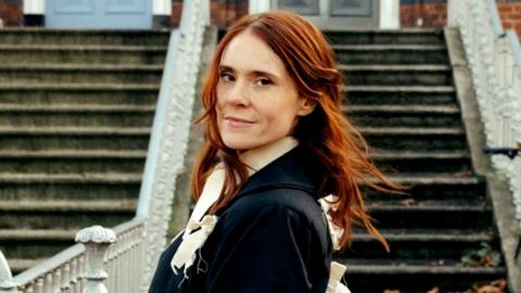 A woman with ginger hair, in a black coat, pictured against some steps