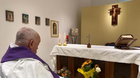 The pope sitting in a wheelchair looking at an altar in a white painted room. There's a bible, Crucifix and some candles on the altar, and a depiction of Christ on the cross hanging above it.