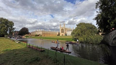 Five punts making their way along the River Cam, in front of Cambridge University colleges, with greenery either side of the river, under a cloudy sky.