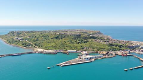 The Isle of Portland in birds-eye view from the sea, showing the port and greenery of the island, surrounded by the sea. 