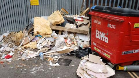 A huge pile of rubbish on the ground next to a red industrial general waste bin