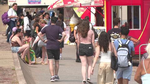 The image shows a busy seaside promenade on a warm day, with large numbers of people walking and gathering outside food kiosks and takeaway stands. A bright red snack kiosk dominates the right side of the picture, while ice cream signs, parasols and groups of people carrying bags and backpacks suggest a popular tourist area during the summer season.