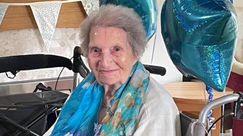 A woman sitting in a wheelchair and smiling to the camera. She has blue birthday balloons attached to her chair and a blue scarf around her neck. She is smiling into the camera and has grey hair.