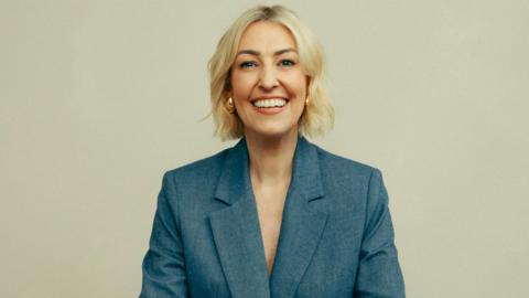 Kelly Cates - a woman with short wavy blonde hair - smiling at the camera and wearing a grey blazer jacket.