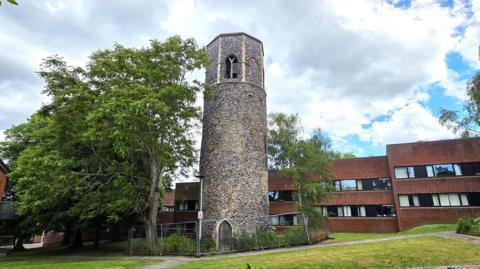 A tall, cylindrical stone tower stands in a grassy courtyard, surrounded by leafy trees and more modern brick buildings with large windows. A fenced area encircles the tower’s base. A partly cloudy sky stretches overhead.