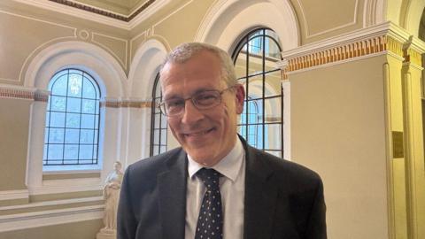 A man with short grey hair, wearing glasses and a suit, pictured from the shoulders up with windows and a statue behind him.