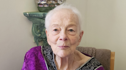 An elderly woman with short white hair and a purple dress.