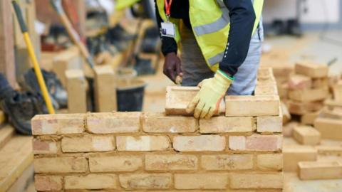 A man in a yellow reflective jacket with yellow gloves on is laying a light brown brick on a small wall. There are other bricks behind, buckets and tools behind him.