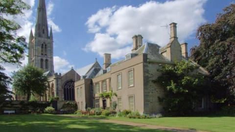 The stone house has a beige render to the front and stands next to a church.  It has tendered lawns to the front with a number of shrubs.