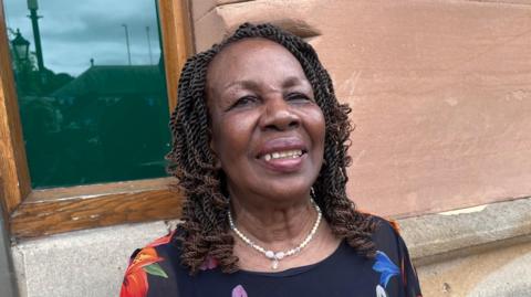Lilian Francis is standing in the middle of the picture looking slightly to the left of the camera. She is wearing a black top with floral patterns on and has a necklace made of white pearls.