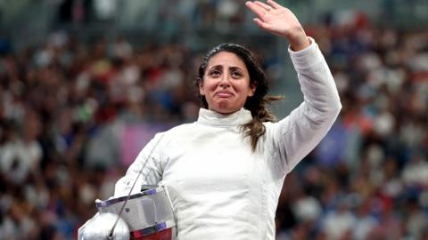 Nada Hafez, wearing a white fencing uniform, holding a sabre in her right hand and cradling a helmet against her hip, looks emotional as she waves to the crowd with her left hand