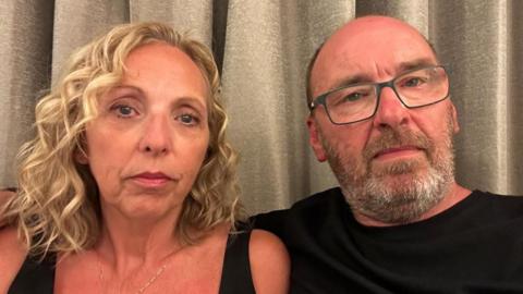 Cynnamon and Tony Church. They have slightly stern looks on their faces. They are both in their 50s. Cynnamon has blonde curly hair and a black vest top on. Tony wears glasses, is balding and has a greying beard. He is wearing a black T-shirt.