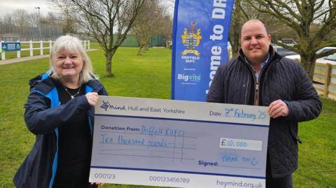 A woman with short blond hair accepts a large cheque for £10,000 from a man with brown hair. Both are smiling and wearing waterproof coats. They are standing in front of a rural rugby ground,