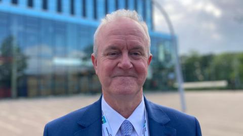 A man in a blue suit with a North Northamptonshire Council lanyard stands in front of the Corby Cube.