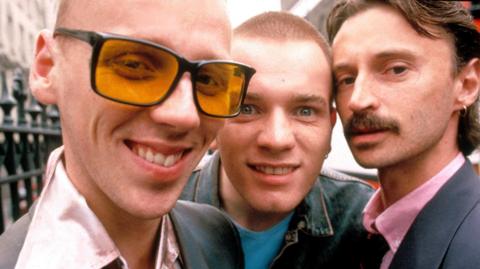 Close up shot from Trainspotting film showing Spud in sunglasses, smiling with pink shirt (Ewen Bremner), Renton sneering in blue T shirt (Ewan McGregor) & Begbie with earring and moustache in bright pink shirt (Robert Carlyle)