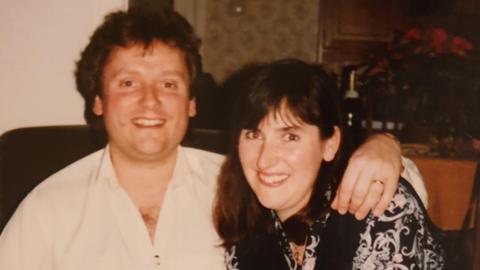 Simon smiles into the camera with his arm around his sister Amanda, who is also smiling. He is wearing a white shirt and has wavy brown hair and a wedding ring on his left hand which rests on Amanda's shoulder. Amanda has long dark brown hair with a fringe and is wearing a black top with a white flower pattern