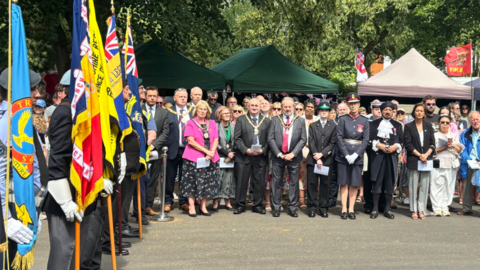 People are holding flags of various colours on the far left of the photo. In the centre and on the right, dozens of people are facing the camera. 