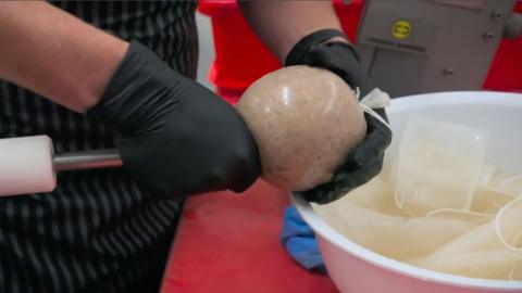 Haggis being piped into a haggis bung. A butcher holds the haggis as it is piped in, wearing black gloves and a black and grey striped apron. There is a white bowl to the right of the image.