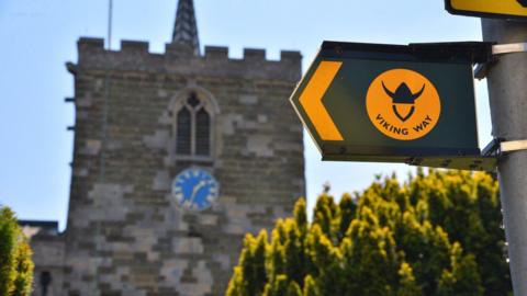 A church can be seen slightly blurred in the background with a blue clock on it and conifers in front of it. A sign can be seen close to the camera with a green and yellow arrow and a yellow circle which says Viking Way with a Viking helmet symbol on it.