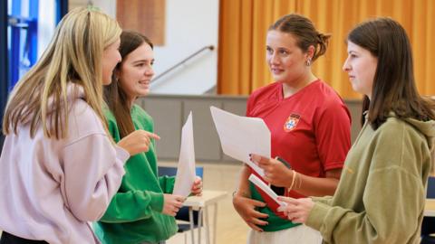 Four young women smile as they compare A-level results. They wear t-shirts and sweatshirts and are standing in their school hall. 