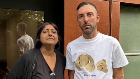 Sarah Singh and Nick Barlow standing side by side outside the entrance to the tower block. Sarah has shoulder length black hair and is wearing a black cardigan. Nick is wearing a white t-shirt with a teddy bear on the front which has had its head removed.