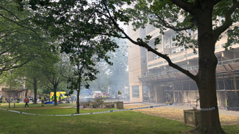 Smoke can be seen in the area with trees surrounding a concrete building which is cordoned off by police tape. 