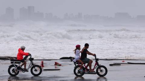 Three people ride two bikes in the coast in Australia with waves crashing in the background