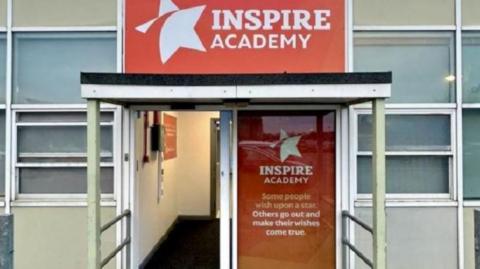 The open doorway of a one-storey building with the words Inspire Academy in large white letters against a red background above the doorway.