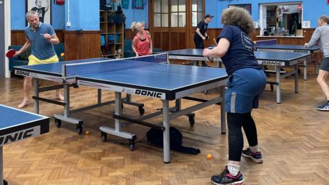 Several people are playing table tennis across multiple tables in an indoor hall with blue walls. Flags and club T‑shirts hang on the walls and orange balls are on the floor.