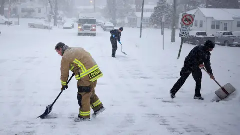 Firefighter and two men in black outfits with shovels clearing snow on road with truck in the background