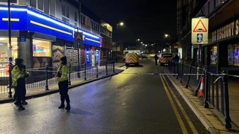 Two police officers stand in front of blue and white police tape in Dewsbury town centre.