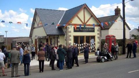 A magnolia building with a wooden-gabled roof, bunting in the windows, with a red post box next to it. A black and gold sign says The Village Shop and the shop is surrounded by villagers including one on a mobility scooter.