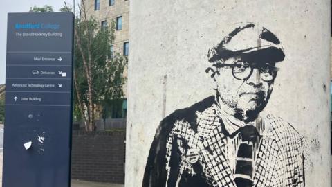 A graffiti image of David Hockney on a pillar with a sign for Bradford College behind