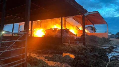 A metal agricultural shed has materials underneath it that are engulfed in bright orange flames