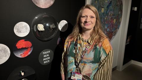 A woman with long light brown hair stands in front of a moon rock exhibition. There are signs on the wall that encourage people to touch part of the exhibition. The woman is wearing a multi-coloured blouse.
