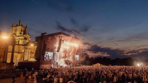 A large crowd in front of a stage with bright lights, and the towers of Blenheim Palace behind.