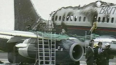 Photograph of the Manchester Air Disaster. This is a still from a BBC News report from August 1985. The charred remains of a large passenger jet are visible, with tome of the roof missing.