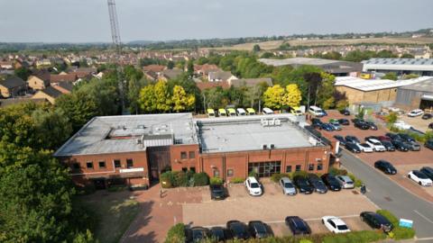 An aerial photo of the East of England Ambulance Service Control room and car park. You can see a radio transmitter at the back of the building, along with a row of ambulances, to the front are a number of cars in the car park. The building is red brick and is two stories high to the left hand side with a flat roof, to the right is a hanger for ambulance parking.