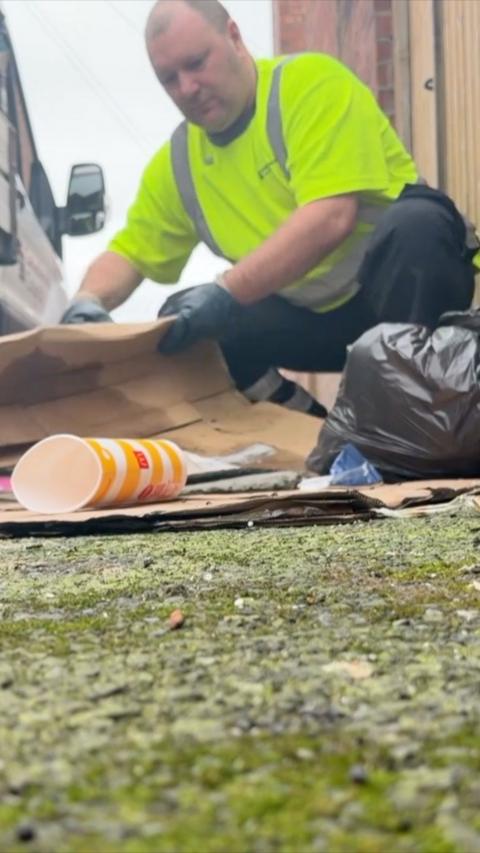 A man in an optic yellow hi-vis t shirt, black trousers and dark blue gloves is crouched down picking up a folded cardboard box. There is a black bag next to his left knee. An empty McDonald's drinking cup is on the left. Mossy pavement takes up a third of the frame from the bottom. 