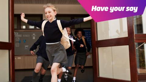 Girl runs out of the school entrance with her arms in the air and a big smile on her face. She is wearing her school uniform and other pupils are following her