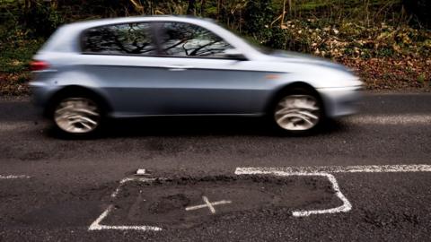 A light blue car driving past a pothole, which has been marked with paint.