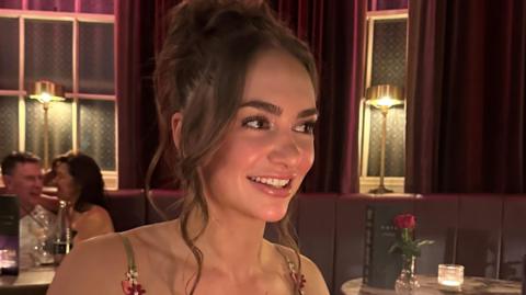 Grace Nevens is sitting in a restaurant and smiling brightly. Her brown hair is pulled up in a ponytail.