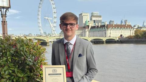 Jack smiles as he holds his award certificate. He stands next to the River Thames with the London Eye wheel and County Hall in the background. He wears dark glasses with a grey tie and suit.